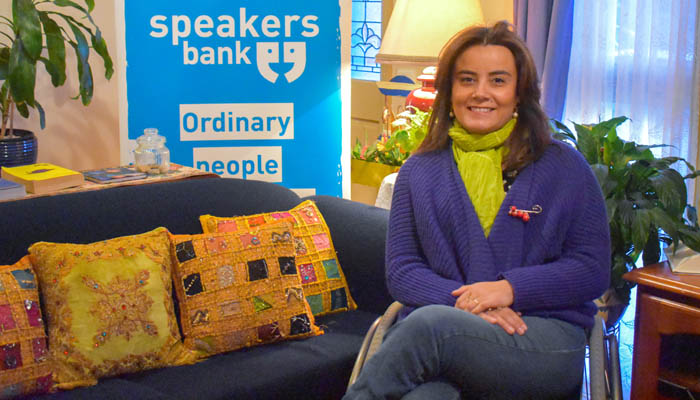 A woman with brown hair in a green turtle neck and purple jacket sits with her hands on her crossed legs. She is smiling at the camera. There is a couch with colourful pillows behind her and a pull up banner that says "Speakers bank - Ordinary people" in blue and white behind her. The rest of the banner we cannot read as it is behind the couch.