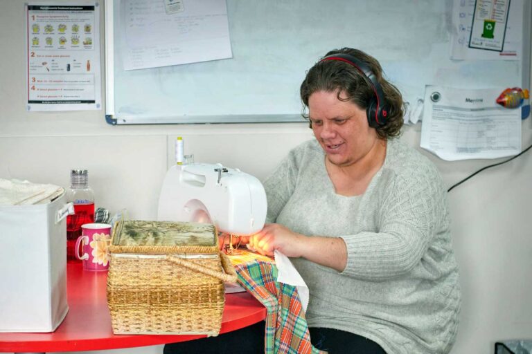 Woman in grey jumper, sewing a green and red colourful blanket. She has black hair and us wearing black headphones. There is a whiteboard behind her and a number of things on the table with the sewing machine, including a coffee cup and a cane basket.