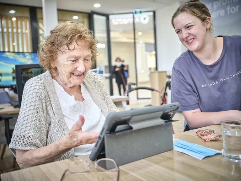 Older woman with curly blonde hair smiles as she looks at an ipad, her hand is waving towards it. A aged care support services woman on the right in a blue t-shirts with their hair pulled back is looking at the iPad, smiling.