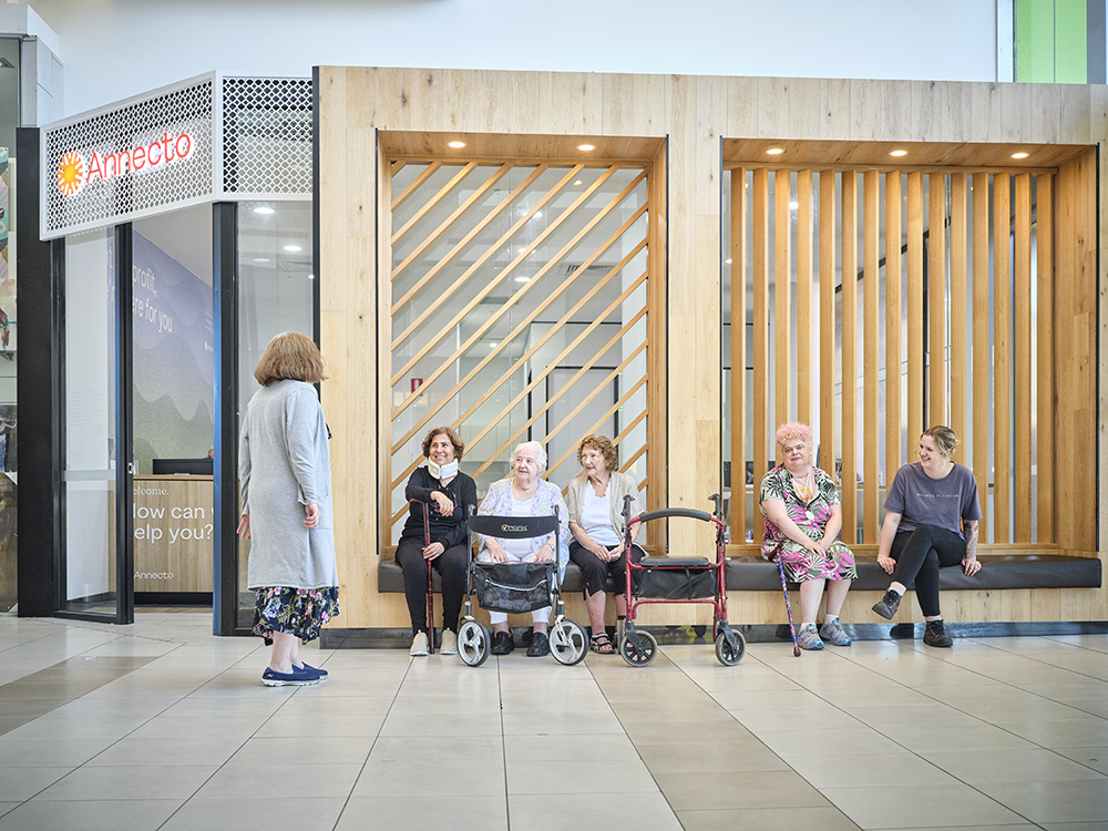 Five women sit in a line on the small ledge at the Annecto Mornington location. They have various mobility devices withthem including a cane and a walker. They are focused on a woman who's standing on the left talking to them.