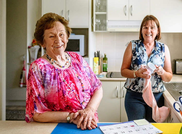 Older woman with brown hair in a pink and purple blouse smiling at camera in a kitchen with some paper and chopping boards in front of her. A younger woman is behind her with brown hair and a floral blue and white top, smiling mid laughter. She is holding a tea cup and red tea towel.