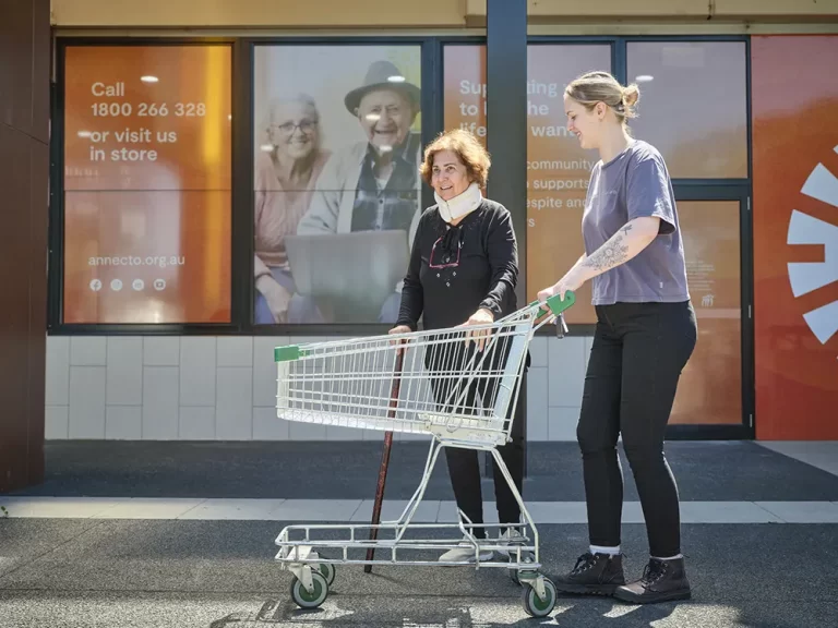 Two women are infront of an Annecto branded store. One, an Annecto volunteer, is walking with a trolley, the other woman beside her, she is holding a cane.