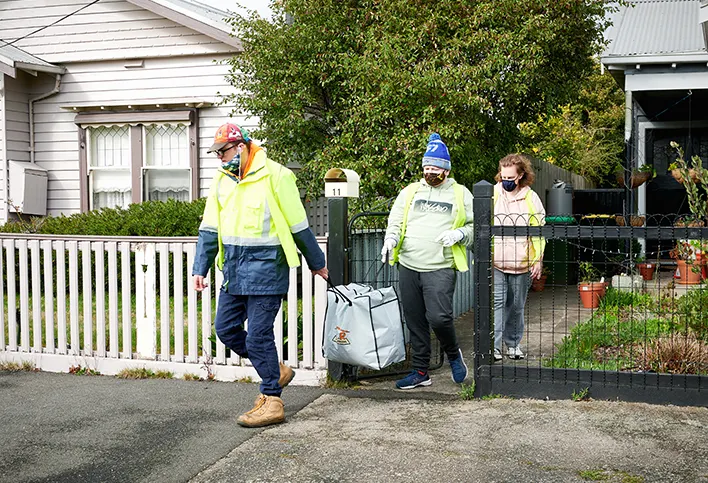 Three people, two men and one woman, are wearing fluro vets over their jumpers and masks of different colours. The men walk in front of her, carrying a large back full of clothes between them. She folows after. They are exiting a grey house to the street, a big white house on the left of them.