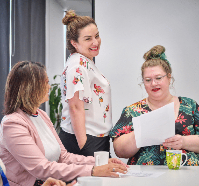Three women in the office, with one standing up and two sitting down, having a laugh together about something ambiguously written on a piece of paper that one of them are holding