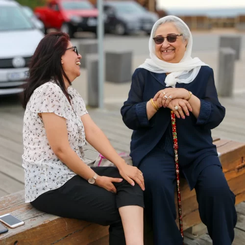 Two women sit on a brick seat, there are cars and concrete posts blurred out in teh background. The two women are talking, not looking at the camera. The woman on the left has black ahir and glasses, she has a a spotted shirt and navy lpants. The woman on the right has black sunglasses, a white hajib and is wearing black clothes. She is leaning her hands on the a colourful walking cane.